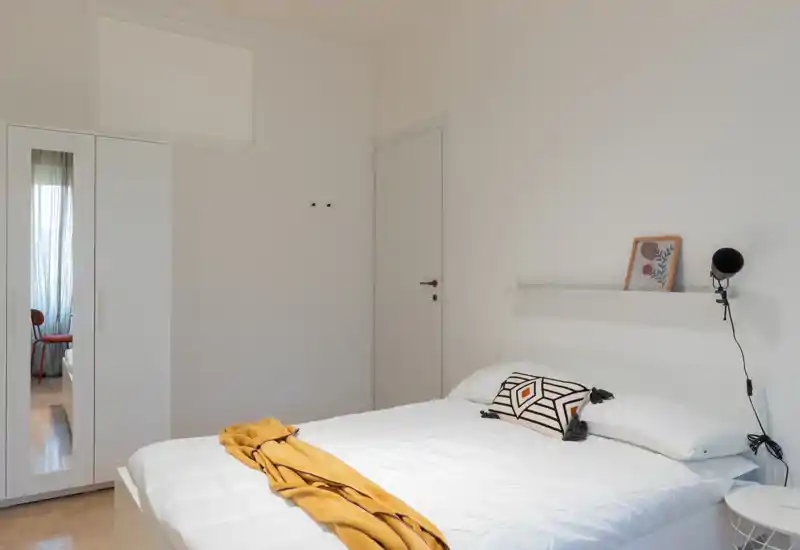 Bright, minimal bedroom featuring a double bed with white bedding, a decorative pillow and a mustard throw; wardrobe with mirrored door and soft natural light.