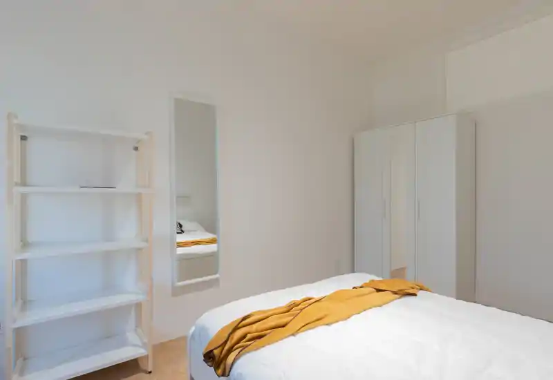 Minimal, bright bedroom with a white bed dressed in a quilt and a mustard throw; wardrobe and full-length mirror add practical storage and depth.
