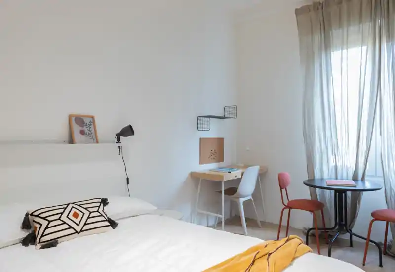 Bright, stylish bedroom with a large bed dressed in white linens, decorative cushions and a cozy mustard throw; includes a small work desk and a round bistro table by the window letting in soft natural light.