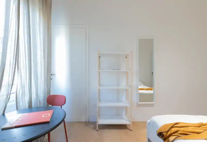 Minimal, bright bedroom corner with a partially visible bed dressed in white and a warm mustard throw; a shelving unit, slim mirror and a small table by a sheer-curtained window complete the scene.