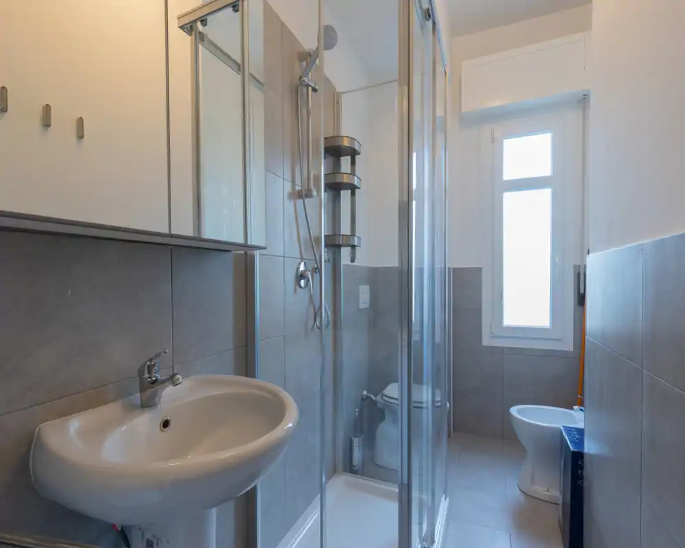 Bright, modern bathroom with a corner glass shower, wall-mounted sink, toilet and bidet; neutral gray tiles and a tall frosted window provide natural light.