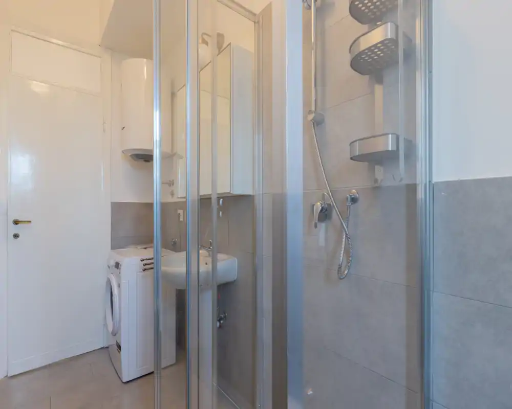 Compact modern bathroom with a glass-enclosed shower, wall-mounted sink and a washing machine. Neutral grey tiles and chrome fixtures give a clean, functional look.
