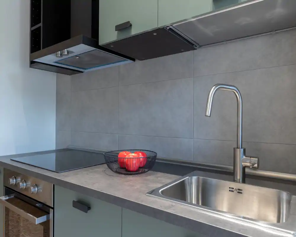 Modern compact kitchen with stainless steel sink, induction cooktop and a decorative bowl of red apples. Clean lines, neutral tiles and soft lighting create a tidy, functional cooking area.