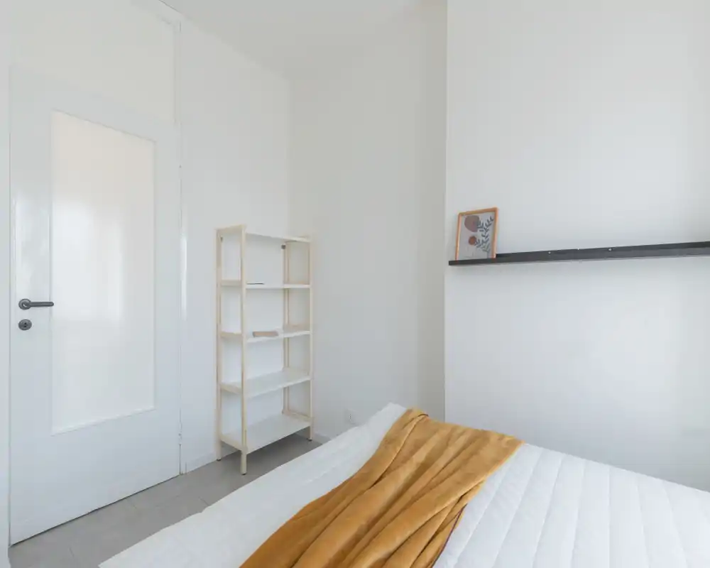 Dormitorio minimalista y luminoso con cama bien hecha, paredes claras y una estantería simple; una manta cálida añade un toque de color.