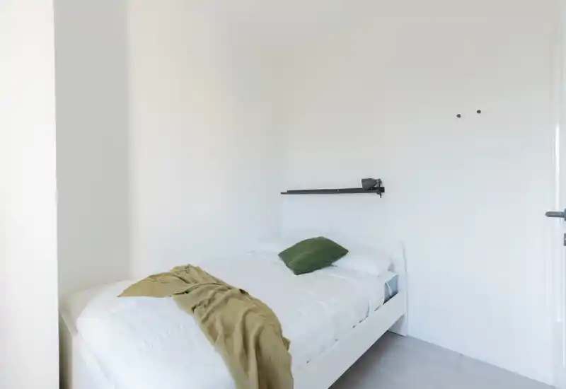 Bright, minimalist single bedroom with a white bed, green pillow and throw. Clean lines and soft natural light make it ideal for a peaceful stay.