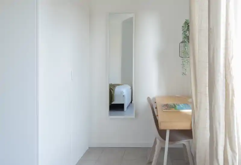 A bright, minimal bedroom corner showing a full-length mirror that reflects a bed; a small wooden desk and chair sit by the window with light curtains and a hanging plant.