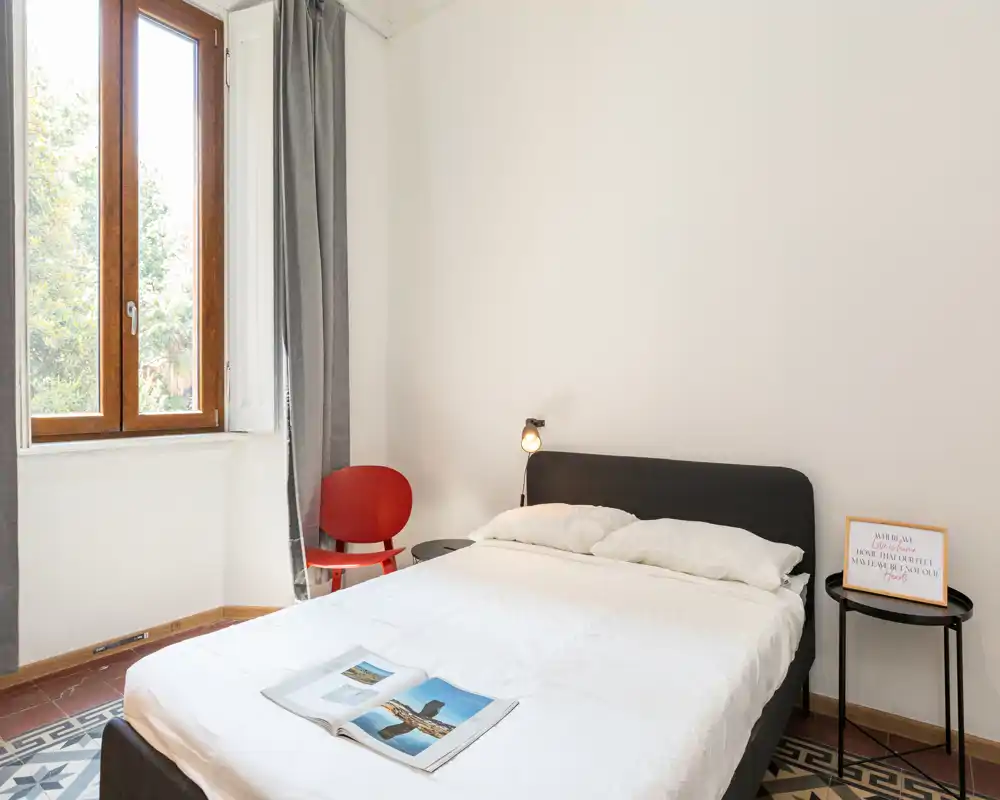 Camera da letto luminosa e minimal con letto matrimoniale, biancheria bianca, lampada da comodino, piccolo tavolino nero e una poltroncina rossa accanto a una finestra alta che lascia entrare luce naturale.