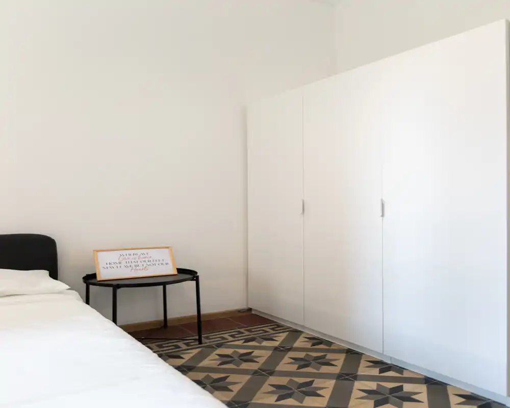 Camera da letto minimal con letto parzialmente visibile, piccolo comodino nero con cornice, grande armadio bianco e pavimento piastrellato decorato — ambiente luminoso, semplice e tranquillo.