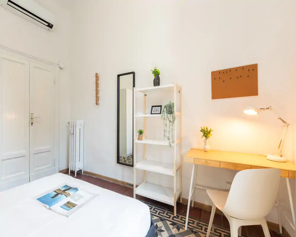 Camera da letto luminosa e minimal con letto ben rifatto, scrivania in legno e scaffale, piante decorative e pavimento con piastrelle a motivi — ideale come spazio accogliente per lavoro e riposo.