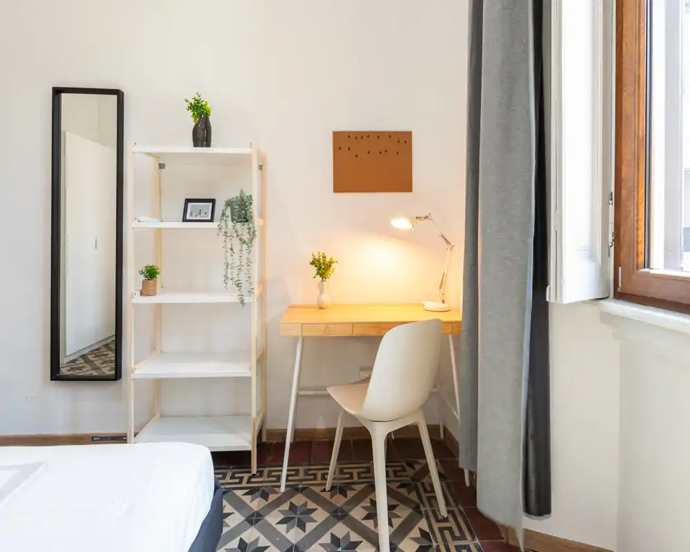 Angolo di camera luminoso e moderno con bordo del letto visibile, scaffalatura e scrivania in legno illuminata vicino alla finestra. Stile pulito e minimal con piante e specchio a figura intera che rendono l'ambiente accogliente.