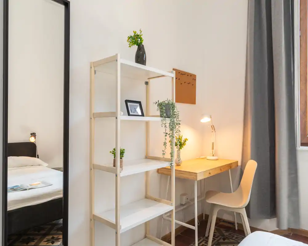 Angolo camera luminoso e ben arredato con letto visibile, una scaffalatura alta e una scrivania in legno compatta con lampada da tavolo — ideale per un’area lavoro accogliente.