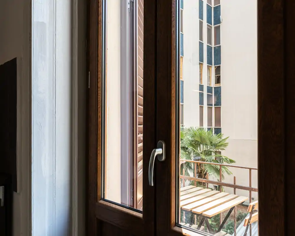 Piccola terrazza soleggiata vista attraverso una finestra con cornice in legno, con tavolo e sedie e vista sugli edifici vicini e una palma.