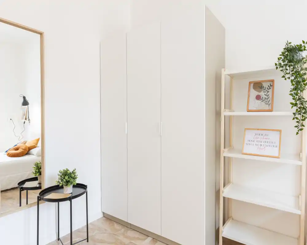 Angolo di camera minimalista con un grande armadio e scaffalatura; uno specchio a sinistra mostra parte del letto con i cuscini, oltre a piante decorative e linee pulite.