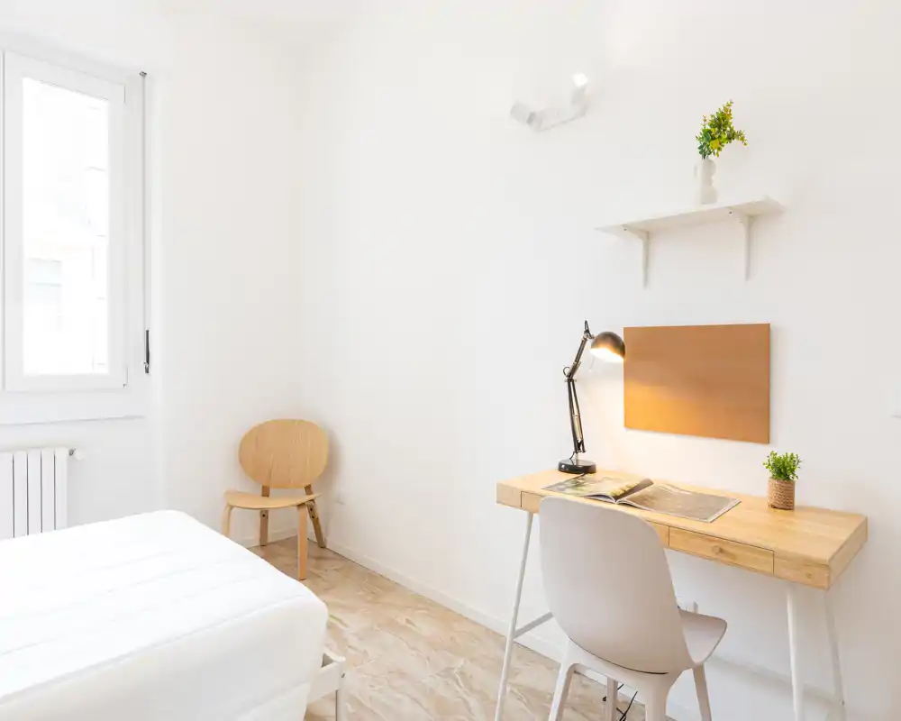 Camera da letto luminosa e minimale con letto bianco, scrivania in legno e sedia. La luce naturale dalla finestra valorizza le linee pulite e l’arredo accogliente.