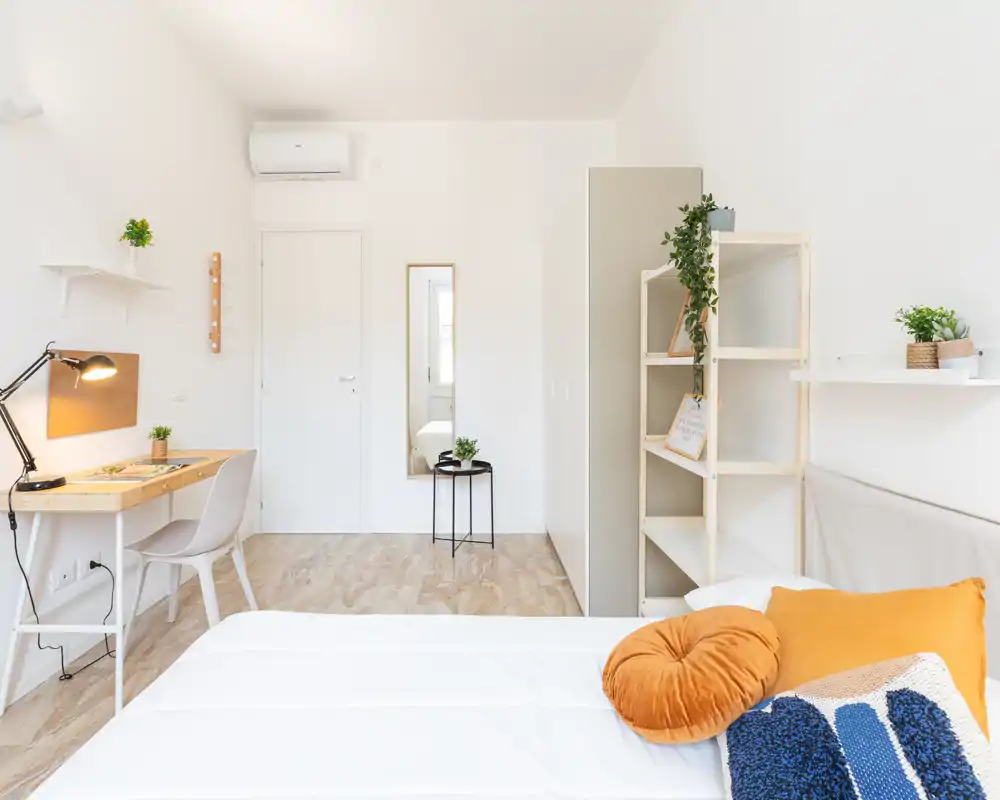 Camera da letto luminosa e minimal con letto rifatto, cuscini caldi e una piccola scrivania: ariosa, moderna e ben arredata per affitti.