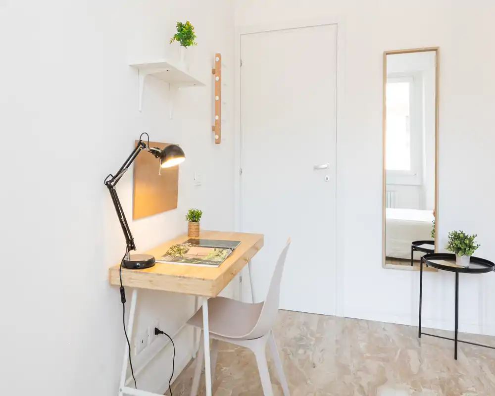 Angolo living luminoso e minimal con una piccola scrivania in legno, lampada da tavolo, sedia e piante decorative. Le pareti bianche e la luce naturale creano un'area di lavoro tranquilla.