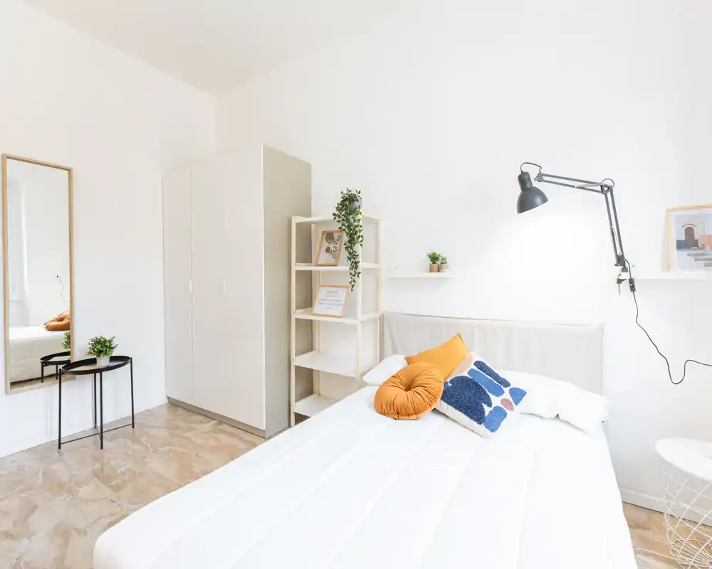 Camera da letto luminosa e minimal con letto matrimoniale ben rifatto e cuscini decorativi, armadio, scaffale e specchio a figura intera—ben illuminata e arredata in modo elegante per annunci.