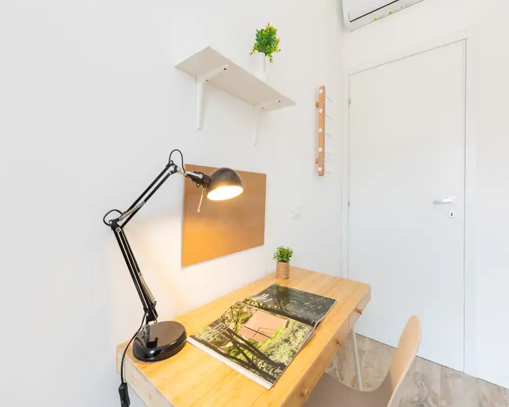 Angolo scrivania compatto e luminoso con piano in legno, lampada da tavolo nera regolabile, mensola a muro e piccola pianta — ideale per lavoro o studio.