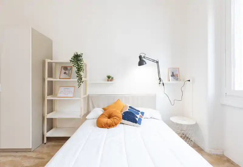 Camera da letto luminosa e minimal con un letto matrimoniale ordinato, cuscini decorativi e una lampada da parete — perfetta per un soggiorno tranquillo e moderno.