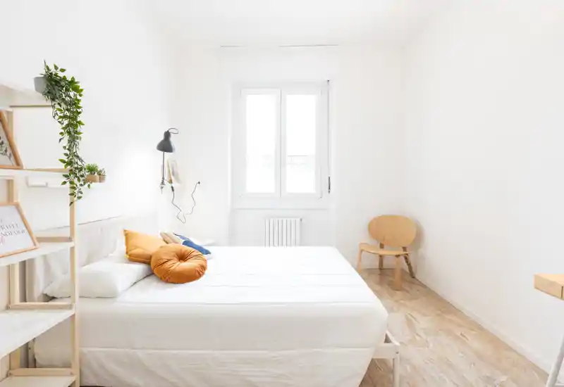 Camera da letto luminosa e minimal con un grande letto bianco, cuscini dai toni caldi e luce naturale dalla finestra — perfetta per annunci moderni.