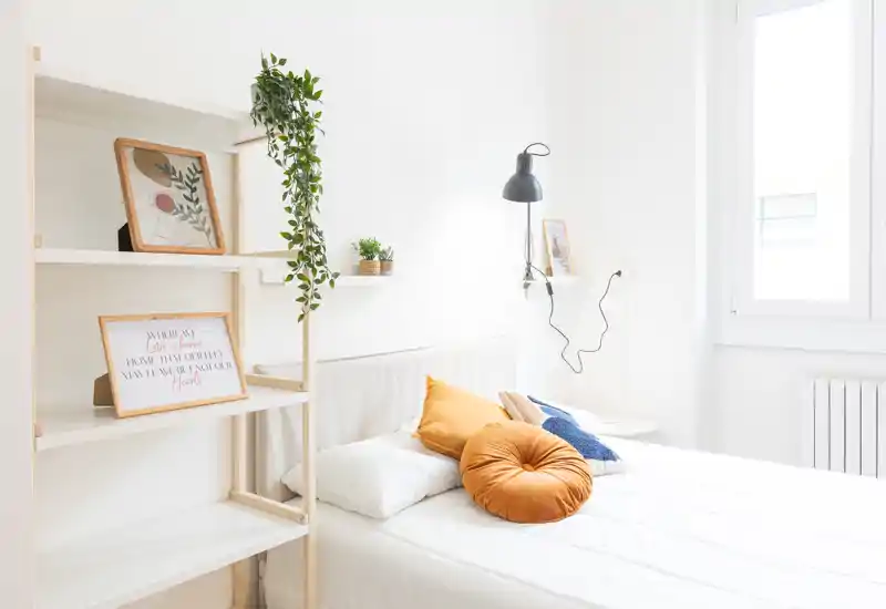 Camera da letto luminosa e minimal con letto centrale, cuscini decorativi color ocra e luce naturale da una finestra ampia; scaffali e piante donano un tocco accogliente.