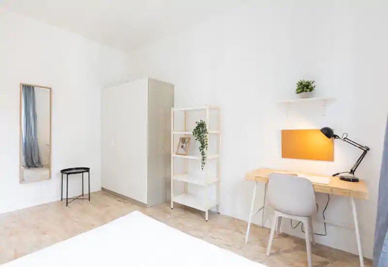 Bright, minimalist bedroom with a white wardrobe, shelving and a wooden desk lit by a warm lamp. The neatly made bed occupies the foreground while the space feels airy and well organized.