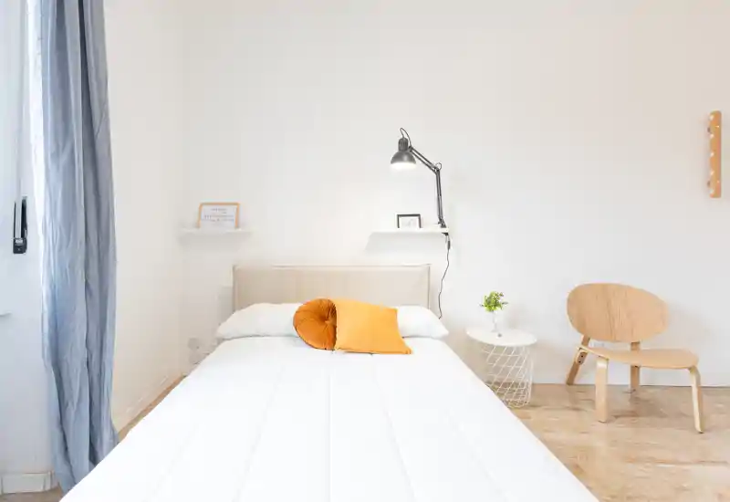 Bright, minimalist bedroom with a large white bed, warm accent pillows, a wall-mounted lamp and simple wooden chair—ideal for modern short-term rentals.