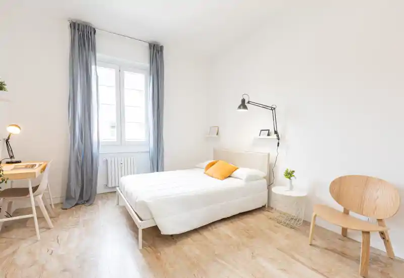 Bright, minimalist bedroom with a full bed, a small desk by the window and light wooden flooring; fresh white linens and a warm accent cushion create an airy, welcoming space.