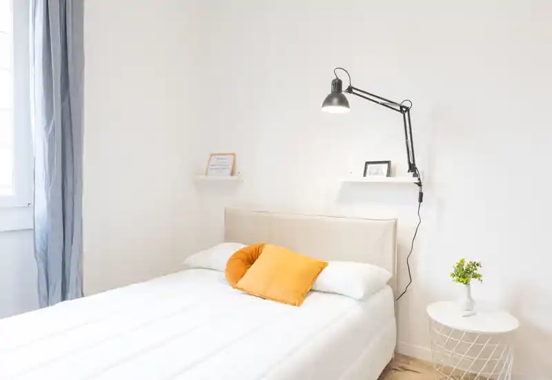 Bright, minimalist bedroom with a neatly made double bed, accented by warm mustard cushions, a wall-mounted adjustable lamp and a small round bedside table with a plant.