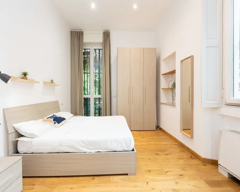 Bright, minimalist bedroom with a double bed, light wood furniture and natural light from a tall window. Clean lines and warm wooden flooring create a welcoming, airy atmosphere ideal for listings.