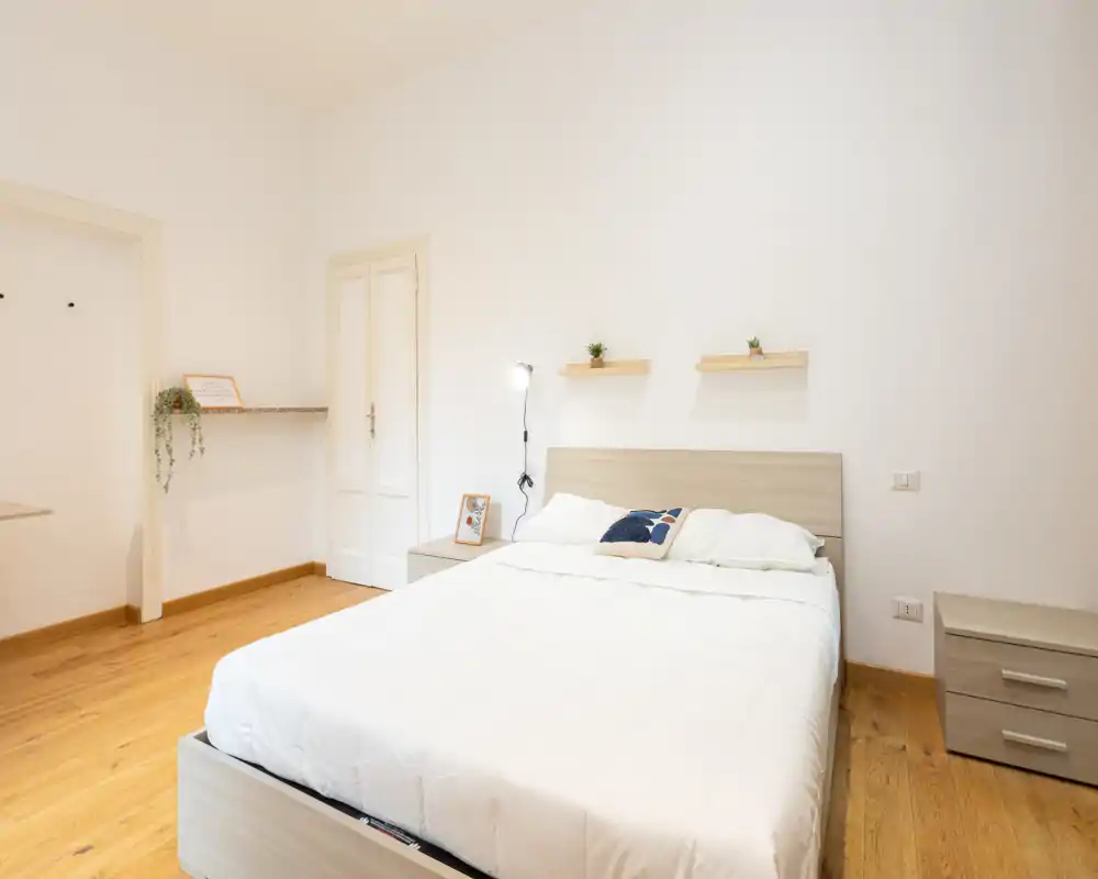 Bright, minimal double bedroom with a wooden floor, neutral decor, bedside tables and simple shelving — ideal for a calm stay.