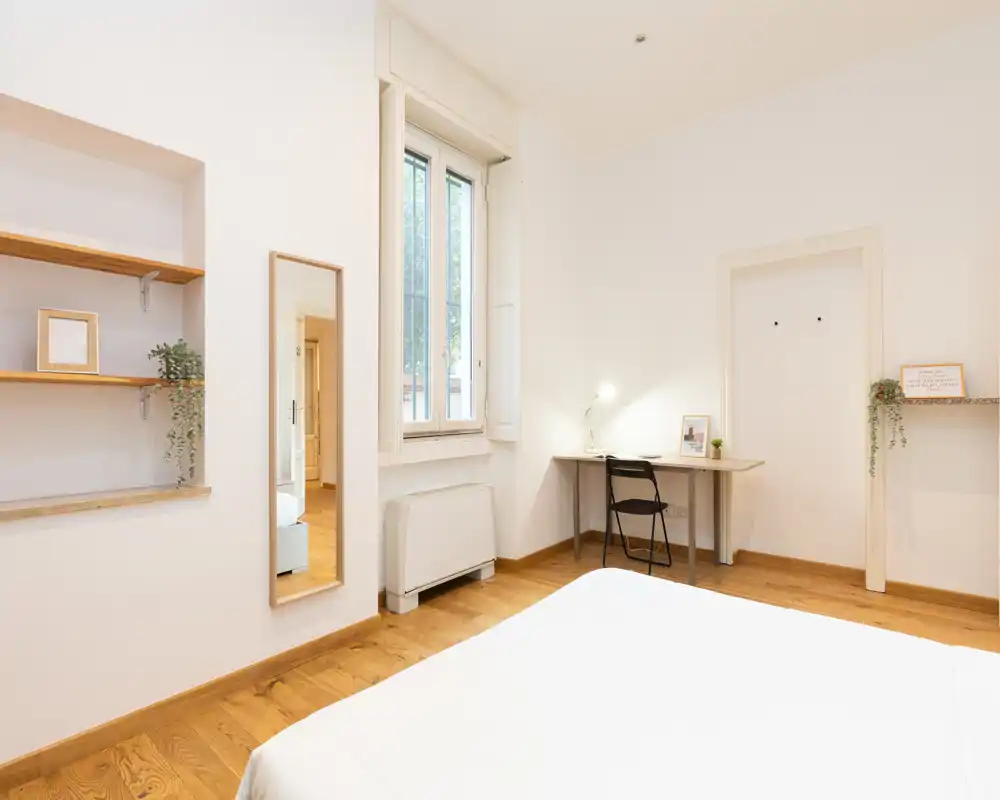 Bright, minimal bedroom with a visible bed, wooden floors, a window providing natural light and a small desk for work or study.