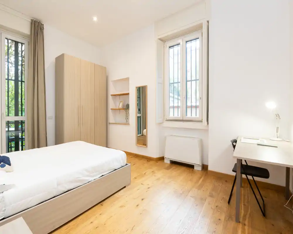 Bright, minimal bedroom with a double bed, wardrobe, desk and natural light from tall windows — ideal for a comfortable stay.