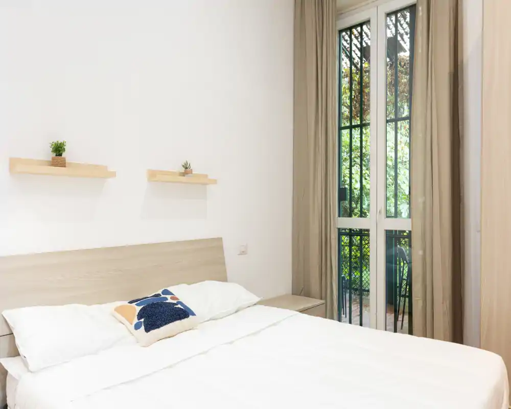 Bright, minimalist bedroom with a double bed, soft linens, small decorative shelves and a large window/door opening to greenery.