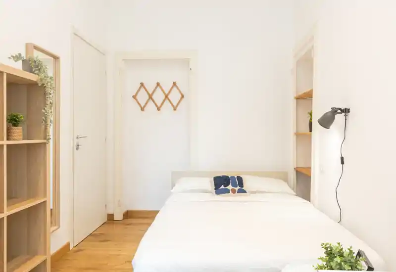 Bright, minimal bedroom with a neatly made double bed, soft linens, wall lamp and simple shelving — a calm, functional sleeping space.