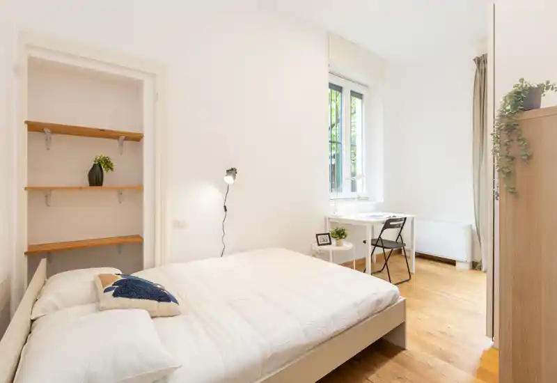 Bright, minimal bedroom with a double bed, bedside light and a small workspace by the window. Natural light and wooden floors create a calm, airy atmosphere ideal for short-term rentals.