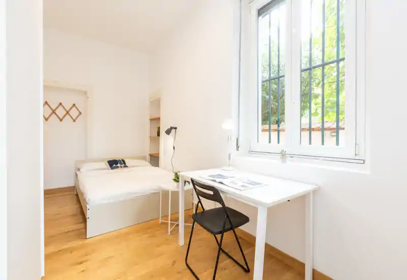 Bright, minimal bedroom with a full bed, a simple desk by the window and warm wooden floors — ideal for a single occupant or short stays.