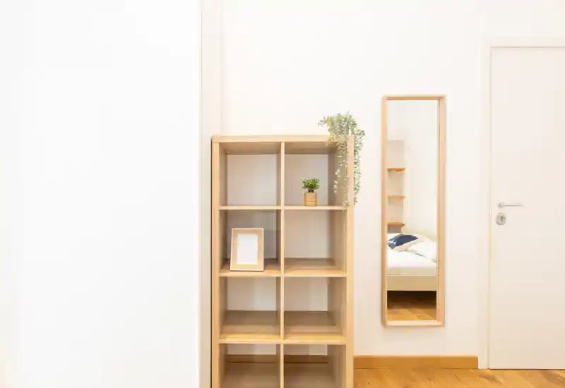 Minimal, bright bedroom corner shot showing a wooden shelving unit and a full-length mirror reflecting part of a bed; clean Scandinavian-style flooring and small plants add a calm, tidy feel.