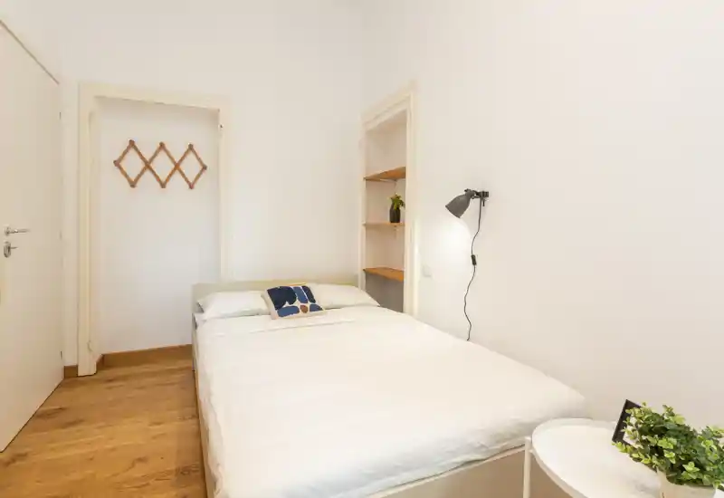 Bright, minimal bedroom with a double bed, wooden floor and simple wall lamp. The room is clean and well-lit with a small bedside table and decorative cushion.