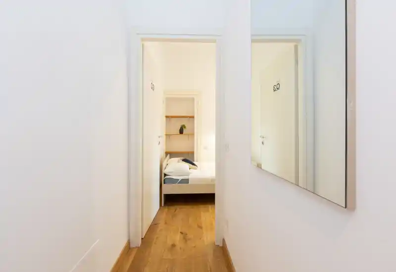 Bright, minimal corridor view leading into a bedroom with a neatly made bed visible through the doorway; simple wooden floor and a large wall mirror in the hall give a clean, modern feel.