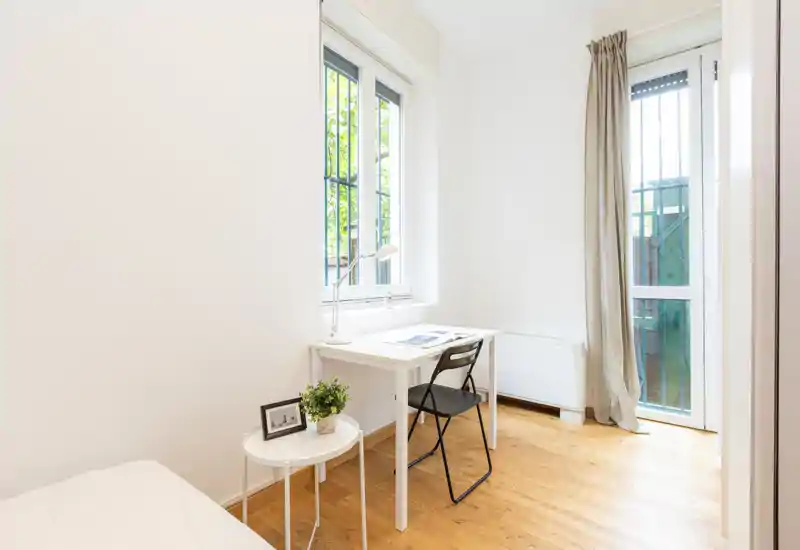 Bright, minimal bedroom corner with a white desk by a window and a small bedside table; natural light and wooden floors create a clean, airy feel.