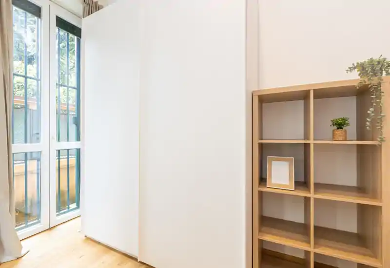 Bright, minimal living area with a large glass door, white sliding wardrobe and wooden cubed shelving accented by small plants — airy and well-lit.