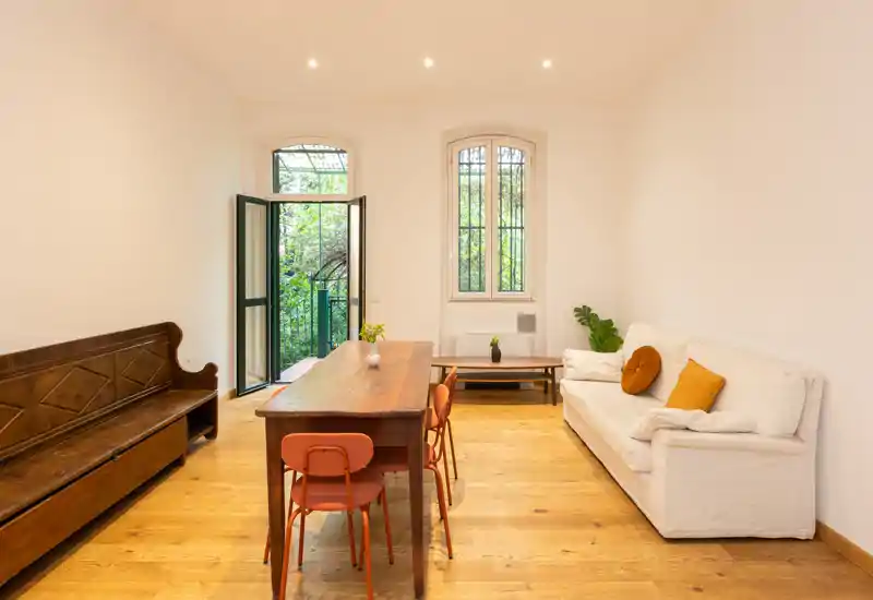 Bright, minimalist living room with a wooden dining table, bench seating and a white sofa with accent cushions; large windows and an open door lead to greenery outside.
