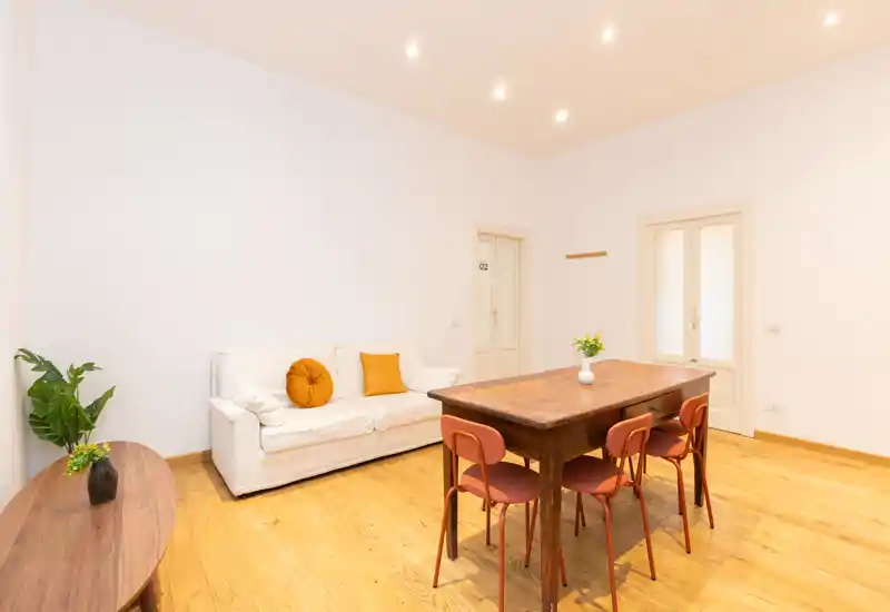 Bright, minimal living area with a white sofa, warm wooden dining table and four chairs — cozy modern styling with simple decor and plants.