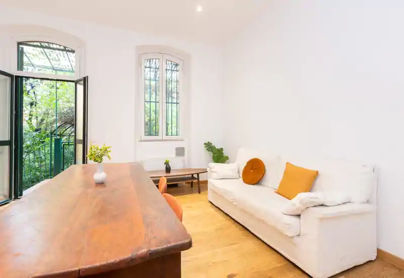 Bright, minimal living area with a white slipcovered sofa, warm wooden table and open window leading to leafy greenery — ideal for relaxed stays.