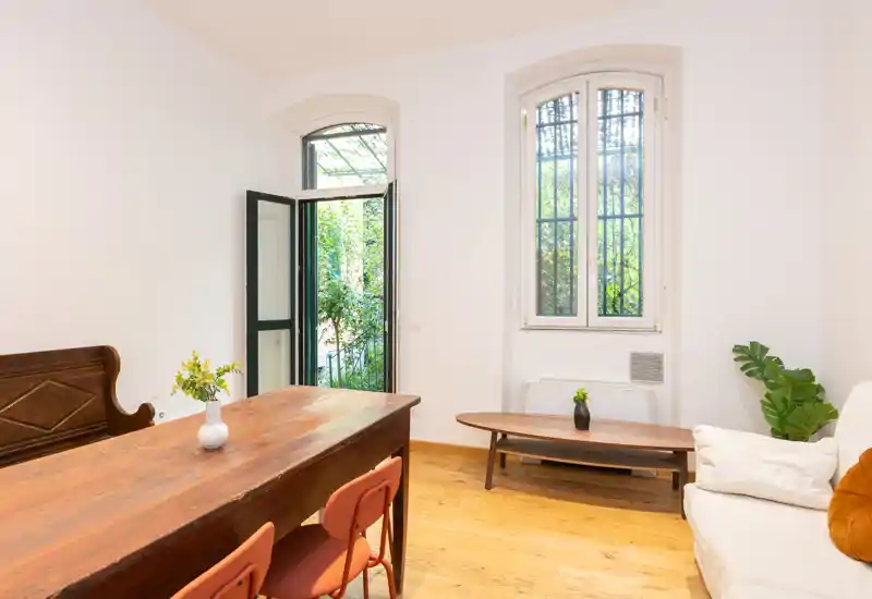 Bright, airy living area with a long wooden table, comfortable sofa and natural light from tall windows and an open door to a small garden.