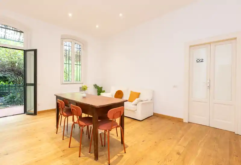 Bright, airy living area with a wooden dining table, four chairs and a small white sofa near a tall window and open door to a garden — ideal for casual dining and relaxing.