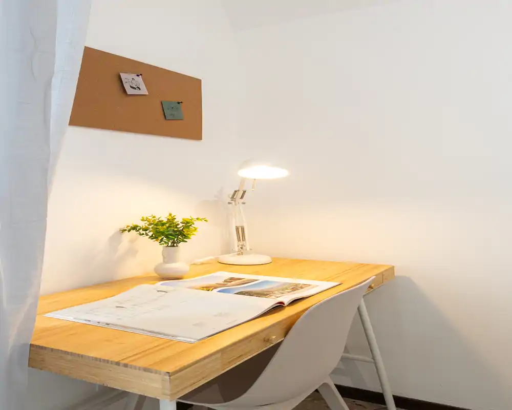 A tidy study nook featuring a wooden desk with an open book, a small potted plant and a lit desk lamp against a plain white wall and corkboard — ideal for work or reading.