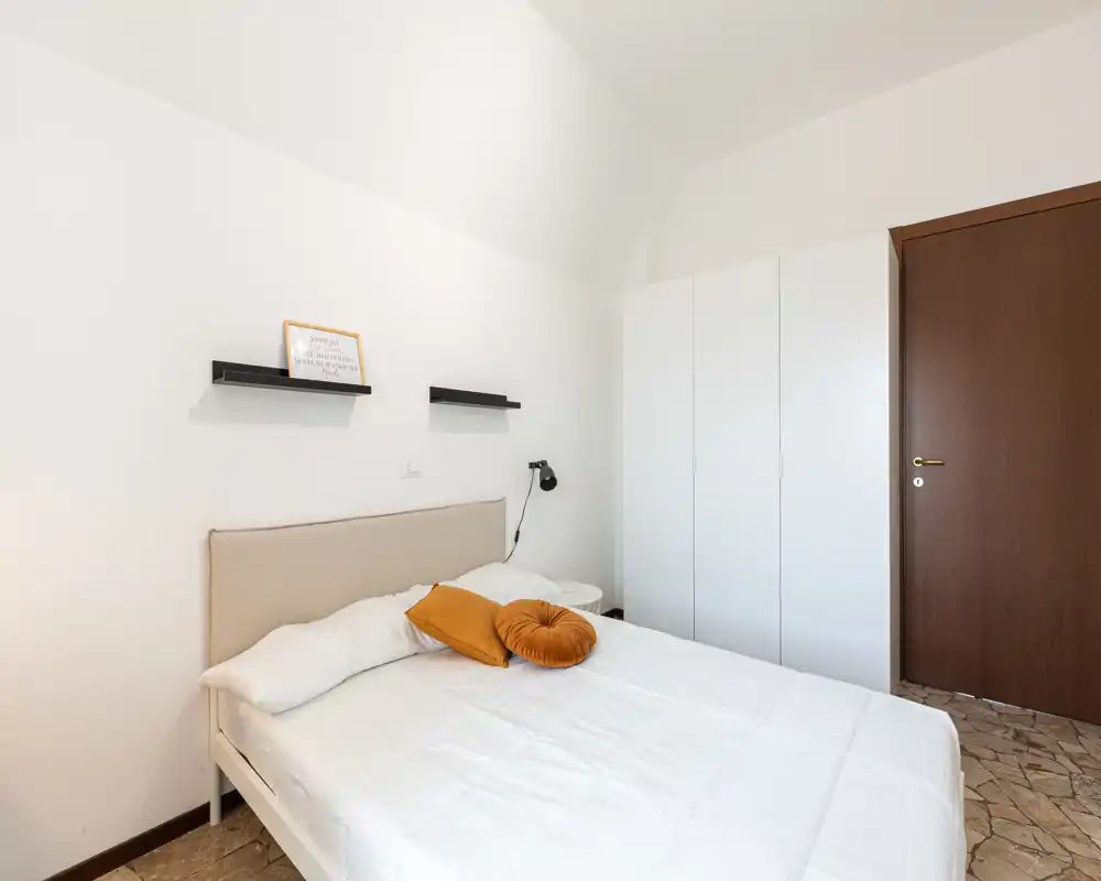 Bright, minimalist bedroom with a neatly made double bed, warm accent cushions and a white wardrobe near the door; clean lines and neutral tones create a calm atmosphere.