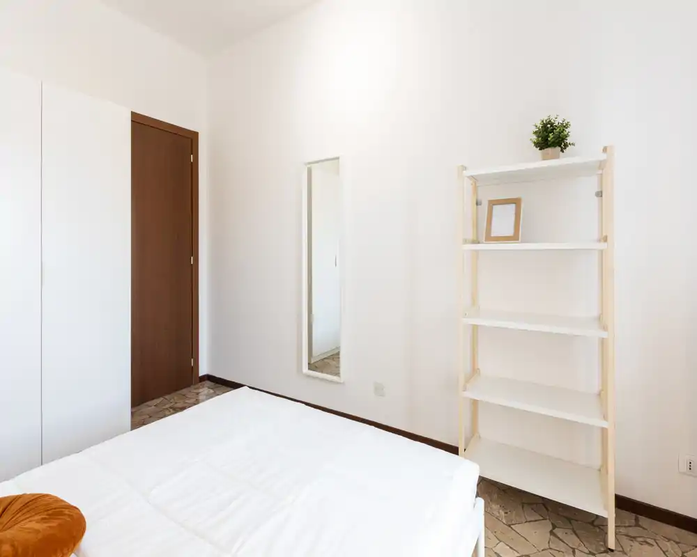 Bright, minimalist bedroom with a white bedspread, simple shelving and a full-length mirror; neutral tones and clean lines create a calm, airy space.